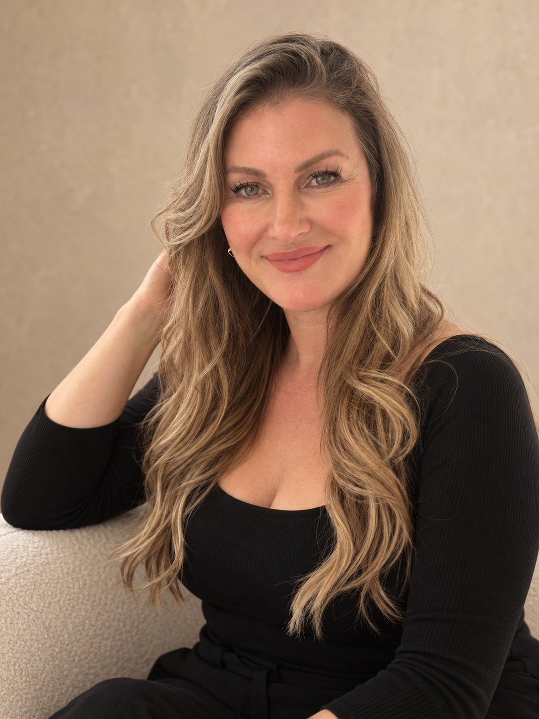 Frame Lash beauty professional with long, wavy blonde hair wearing a black top against a warm beige studio background.
