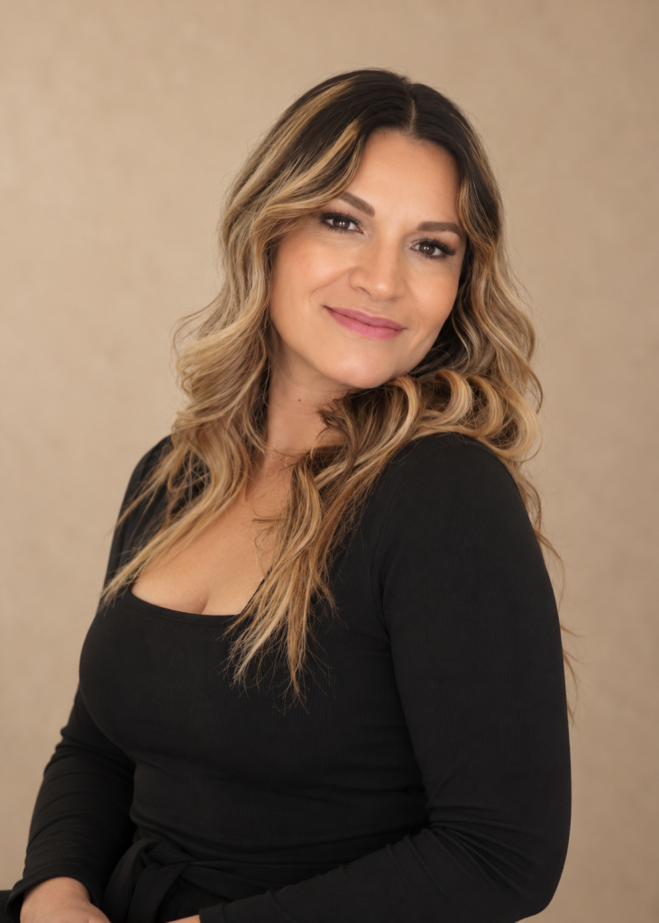 Frame Lash team member with highlighted wavy hair wearing a black top against a warm beige studio background.